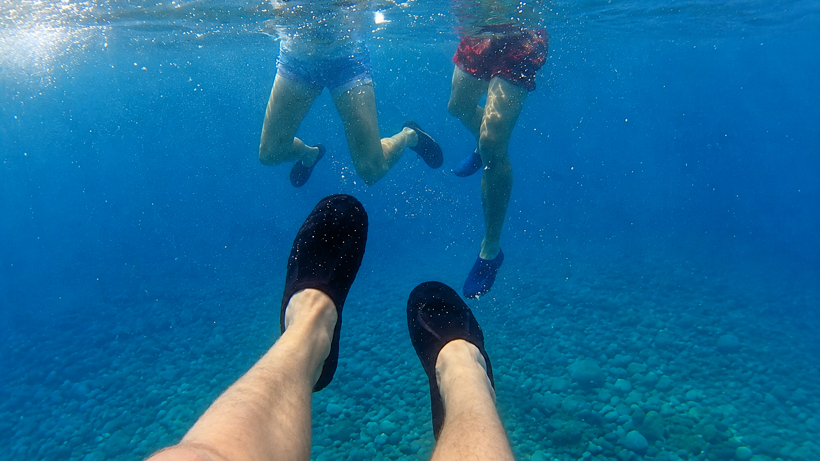 Underwater view of swimmers in water shoes above a pebble seabed